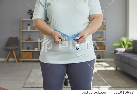 Close up cropped photo of a fat overweight woman measure the waist with measuring tape at home. 131010658