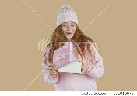 Amazed redhead girl with curly hair opening a present gift box and smiling on beige background. 131010676