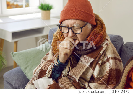 Sick senior man in hat wrapped in a blanket suffering from seasonal flu or cold at home. 131012472