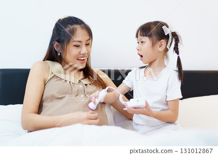 Lovely Asian mother and daughter relaxing - lying down on a bed in bedroom. A girl is excited to listen sound of the baby inside her mother stomach. Lovely Asian mother and daughter relaxing - lying down on a bed in bedroom. A girl is excited to listen sound of the baby inside her mother stomach. 131012867
