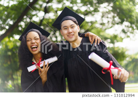A group of diverse ethnicity graduated students enjoy their graduation celebrating together in a campus. Asian male and African female student taking a picture together in their graduation ceremony. A group of diverse ethnicity graduated students enjoy their graduation celebrating together in a campus. Asian male and African female student taking a picture together in their graduation ceremony. 131012994