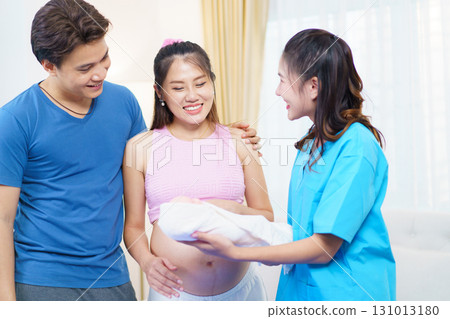 An Asian husband and his pregnant wife are learning how to bathe a newborn baby, guided by a nanny nurse who is demonstrating the bathing process using a baby doll. 131013180