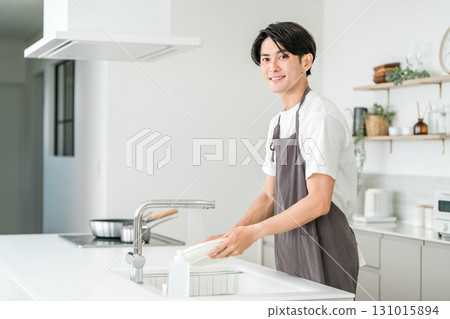 Young Asian man in an apron washing dishes in the kitchen Young Asian man in an apron washing dishes in the kitchen 131015894