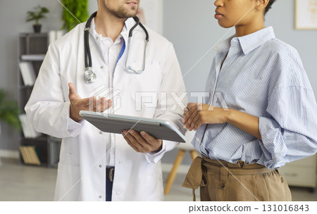 Young man doctor showing test results on tablet to a sick woman patient standing in medical office Young man doctor showing test results on tablet to a sick woman patient standing in medical office 131016843