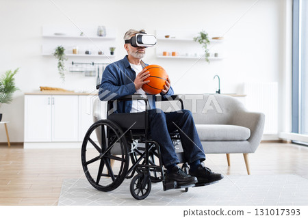 Senior man sitting in wheelchair indoors wears VR headset holding basketball, enjoying technology. Interior setting includes sofa, shelves. Emphasizes virtual reality, accessibility 131017393