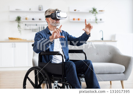 Senior man wearing VR headset explores virtual interface while seated in wheelchair indoors. Concept of accessibility, modern technology, and gaming for older adults. 131017395
