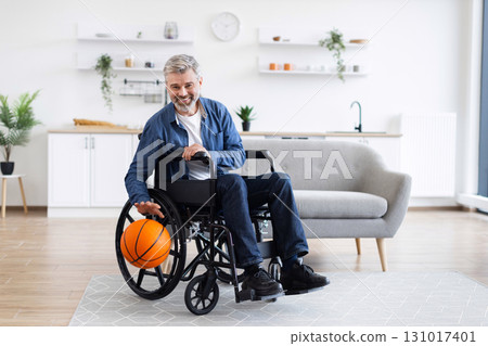Gray-haired man wearing denim and white shirt in wheelchair practicing with basketball, indoors. Gray-haired man wearing denim and white shirt in wheelchair practicing with basketball, indoors. 131017401