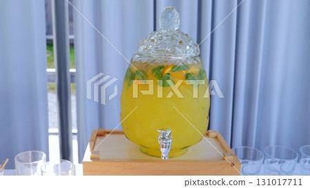 Glass beverage dispenser with yellow lemonade, orange slices and mint leaves, placed on a wooden crate, perfect for summer parties or gatherings Glass beverage dispenser with yellow lemonade, orange slices and mint leaves, placed on a wooden crate, perfect for summer parties or gatherings 131017711