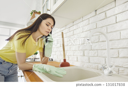 Worried housewoman sing plunger to unclog sink in kitchen using smartphone to call plumbing service 131017838