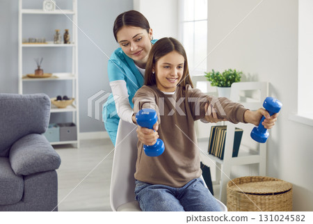 Child girl doing exercise using dumbbells with support from a young friendly female nurse. 131024582