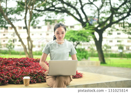 Pretty young woman sitting in park working on laptop remote job Pretty young woman sitting in park working on laptop remote job 131025814
