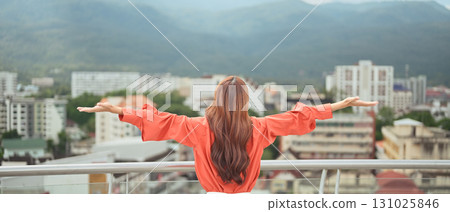 A businesswoman on a rooftop terrace, arms open, enjoying a refreshing break with city and mountain views A businesswoman on a rooftop terrace, arms open, enjoying a refreshing break with city and mountain views 131025846