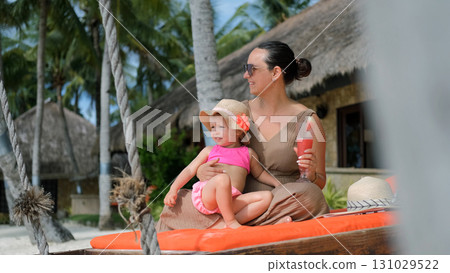 Young woman relaxes on a sunbed with her little daughter at tropical beach enjoying a watermelon shake under the sun Young woman relaxes on a sunbed with her little daughter at tropical beach enjoying a watermelon shake under the sun 131029522