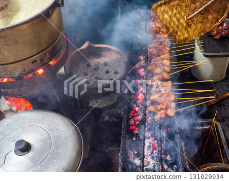 Street food vendor grilling smoky meat skewers over charcoal, using a woven fan to control flame. Vibrant, authentic culinary scene with dynamic motion and texture. Street food vendor grilling smoky meat skewers over charcoal, using a woven fan to control flame. Vibrant, authentic culinary scene with dynamic motion and texture. 131029934