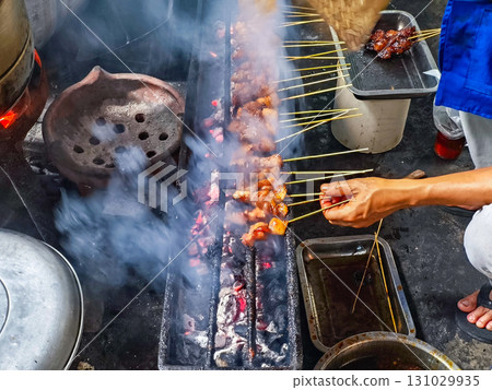 Street food vendor grilling smoky meat skewers over charcoal, using a woven fan to control flame. Vibrant, authentic culinary scene with dynamic motion and texture. Street food vendor grilling smoky meat skewers over charcoal, using a woven fan to control flame. Vibrant, authentic culinary scene with dynamic motion and texture. 131029935