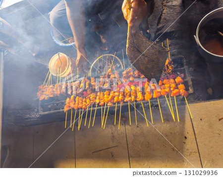 Street food vendor grilling smoky meat skewers over charcoal, using a woven fan to control flame. Vibrant, authentic culinary scene with dynamic motion and texture. Street food vendor grilling smoky meat skewers over charcoal, using a woven fan to control flame. Vibrant, authentic culinary scene with dynamic motion and texture. 131029936