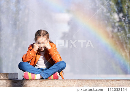 Smiling caucasian female child sitting by fountain with rainbow in bright Smiling caucasian female child sitting by fountain with rainbow in bright 131032134