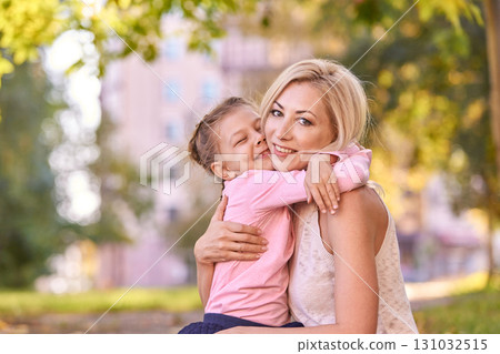 Caucasian adult female and child embrace in sunny park with greenery and urban 131032515
