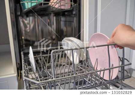 Adult female hand loading dirty dishes in dishwasher open door, pink plates 131032829