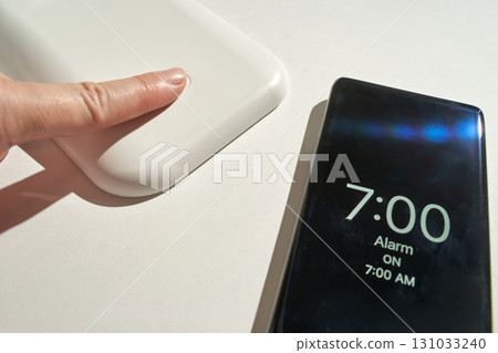 Female hand touching white wireless charger near smartphone with 7:00 am alarm 131033240