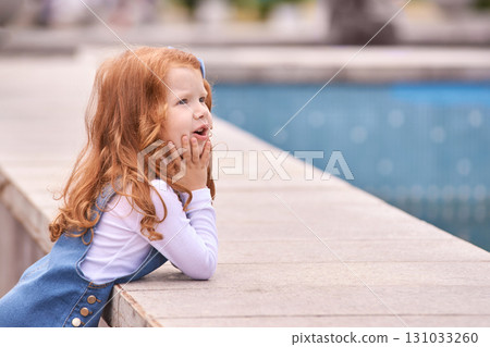 Curious caucasian young girl with red hair contemplating near a pool in outdoor 131033260