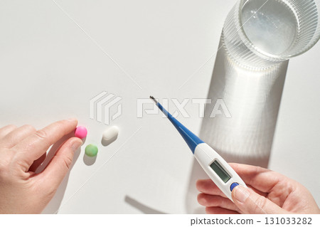 Hands holding pills and digital thermometer on white surface with glass Hands holding pills and digital thermometer on white surface with glass 131033282