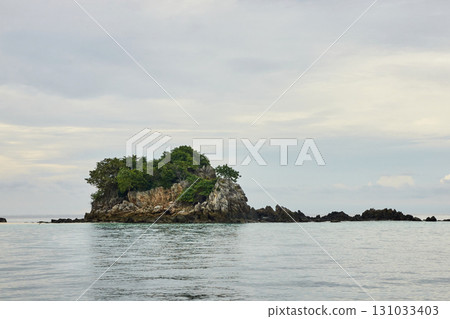 Tranquil small rocky island with lush greenery in calm ocean waters under Tranquil small rocky island with lush greenery in calm ocean waters under 131033403