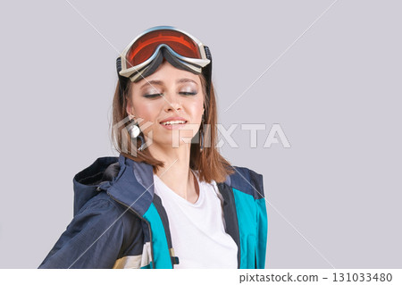 Young caucasian female skier in blue jacket with ski goggles relaxing on gray Young caucasian female skier in blue jacket with ski goggles relaxing on gray 131033480