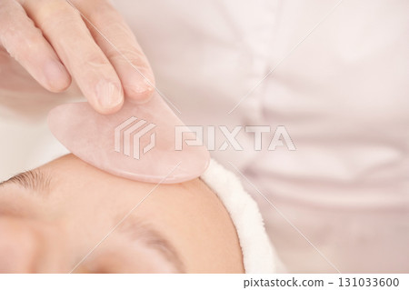 Caucasian female receiving gua sha facial massage with rose quartz stone in spa 131033600