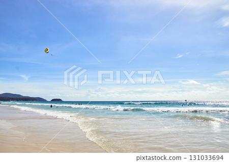 Sunny beach with clear blue skies and waves as people enjoy parasailing 131033694