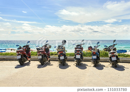 Row of parked scooters facing ocean on sunny beachfront roadside with clear sky Row of parked scooters facing ocean on sunny beachfront roadside with clear sky 131033780