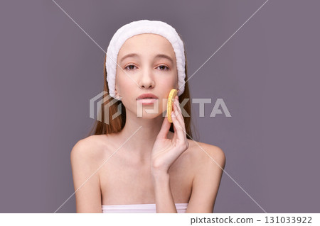 Young caucasian female in makeup headband cleansing face with sponge on neutral 131033922
