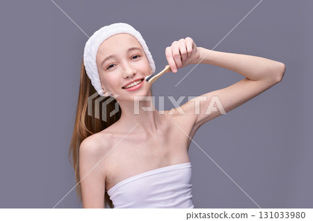 Young caucasian female smiling while applying facial mask with brush on grey Young caucasian female smiling while applying facial mask with brush on grey 131033980
