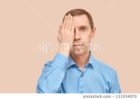 Caucasian young adult male covering one eye with hand in blue shirt against 131034070