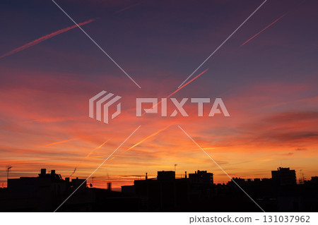 A vibrant sunset with shades of orange, pink, and purple fills the sky. Silhouettes of buildings and contrails are visible against the colorful backdrop. 131037962