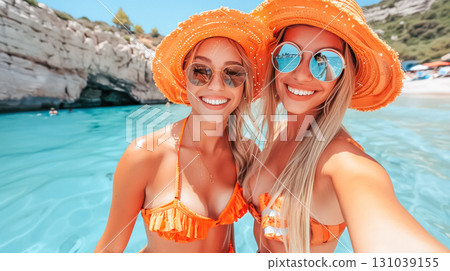 Two smiling women in vibrant orange hats enjoying a sunny day at the beach with clear blue water 131039155