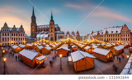A beautiful, wide-angle shot of a traditional German Christmas market set up in a historic town square during the evening. Rows of wooden stalls with snow-dusted roofs are illuminated by glowing light 131041844