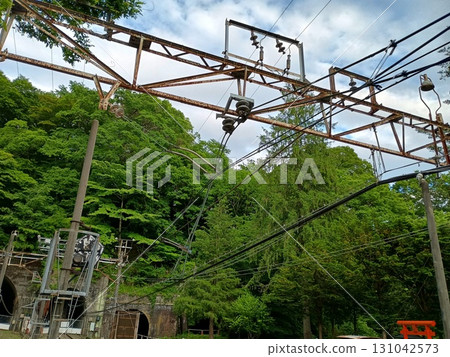 A panoramic view of the railway infrastructure surrounded by forest, with the entrance to a railway tunnel in the mountains and damaged electrification equipment made of rusty overhead power lines. 131042573