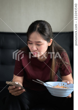 Asian woman sitting at home in sofa eating instant noodles from bowl Asian woman sitting at home in sofa eating instant noodles from bowl 131043540