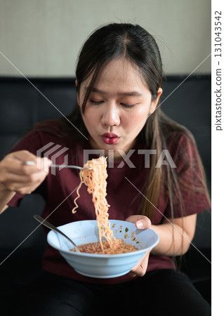 Asian woman sitting at home in sofa eating instant noodles from bowl Asian woman sitting at home in sofa eating instant noodles from bowl 131043542