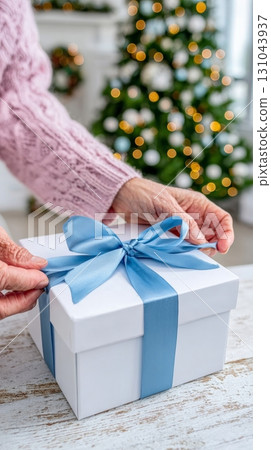 Hands are tying a ribbon around a gift box in front of a decorated Christmas tree 131043937