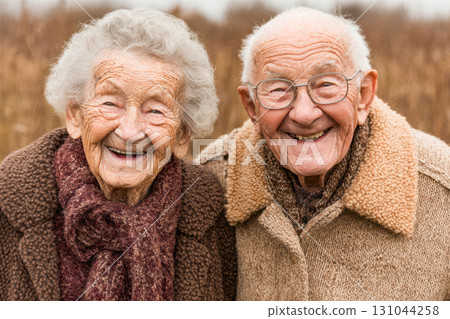 Elderly couple smiling warmly in cozy sweaters against a natural outdoor background 131044258