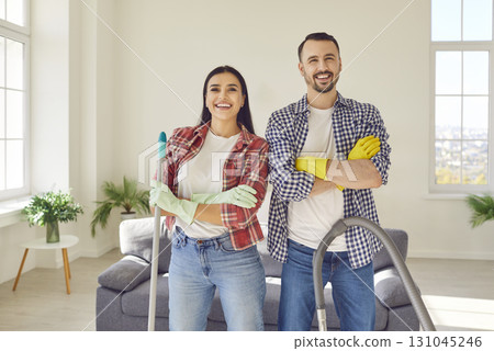 Happy couple standing at home with vacuum cleaner and mop and looking cheerful at camera. 131045246