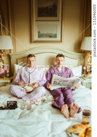 Two men are sitting on a bed, one of them reading a newspaper 131046009