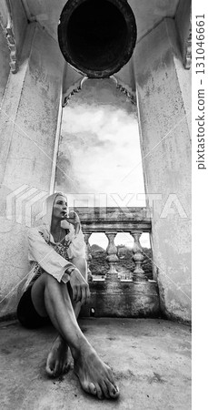 A barefoot woman wearing a white hoodie sits contemplatively on a stone surface. A large structure resembling a bell and an arched window frame a background of clouds and mountains. 131046661