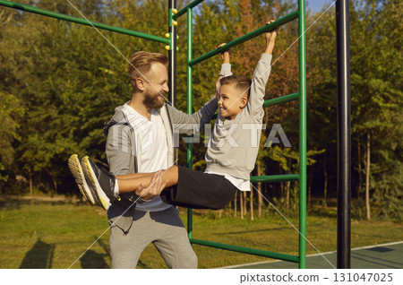 Happy smiling father helping his child boy to do sport fit exercises on gymnastic bar outdoors. 131047025