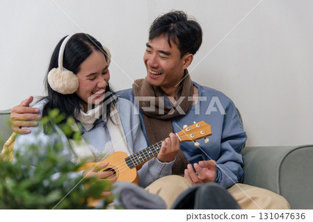 Winter couple shares happy home indoor time, people enjoying cozy moments playing ukulele, man wearing scarf and woman earmuffs for warm day, creating joyful winter memories. 131047636