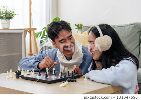 Winter couple, happy people, enjoy indoor day at home, playing lively game of chess, sharing laughter and warmth, cozy winter atmosphere fills living space, creating joyous memories. 131047656