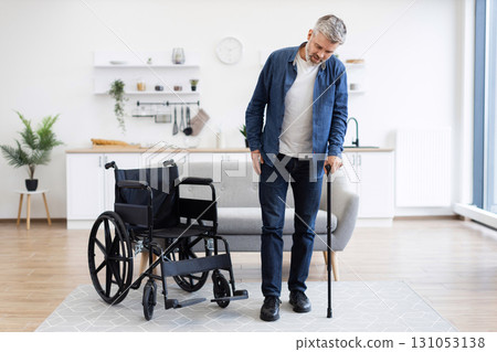 Mature disabled man learning to walk with assistance of cane, standing by wheelchair. Bright modern home interior, independence, recovery. Middle-aged male showing progress in rehabilitation at home. 131053138