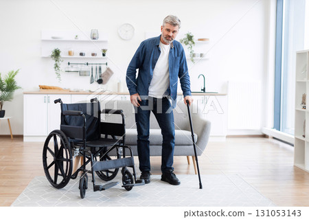 Middle-aged white man with disability using cane in home setting, wheelchair nearby. Depicts determination, recovery, mobility aids, progress within comfortable contemporary setting 131053143